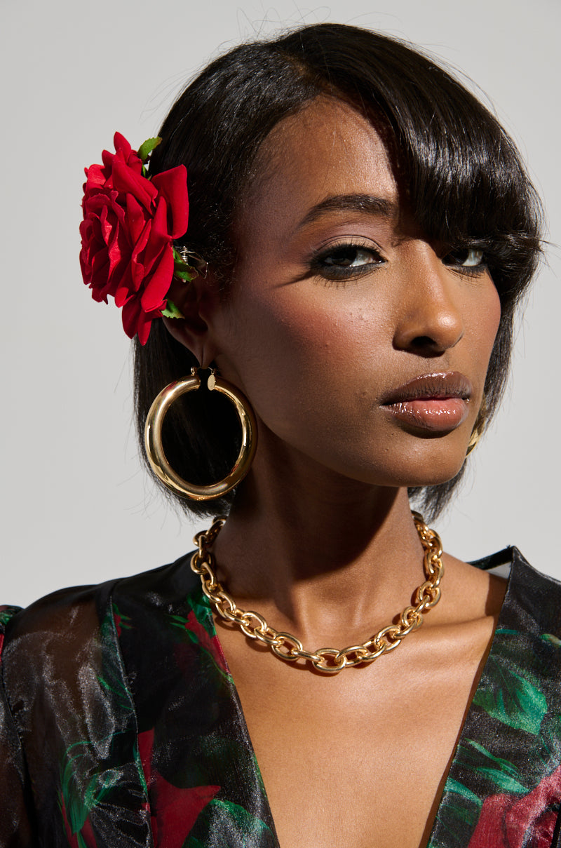 A woman with dark hair, highlighted by a Dolce Vita Rose Hair Clip, adorned in large gold hoop earrings and a gold chain necklace, dons a black floral-patterned top. She poses against a light gray background with soft makeup. 1