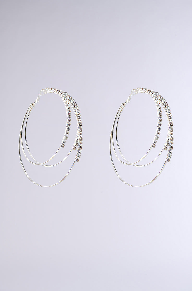 Discover BLING NIGHTS LAYERED HOOP EARRING: elegant silver hoops featuring intertwined smooth metal loops and a rhinestone-embellished loop, set against a pale gray backdrop to add sparkle to any ensemble.