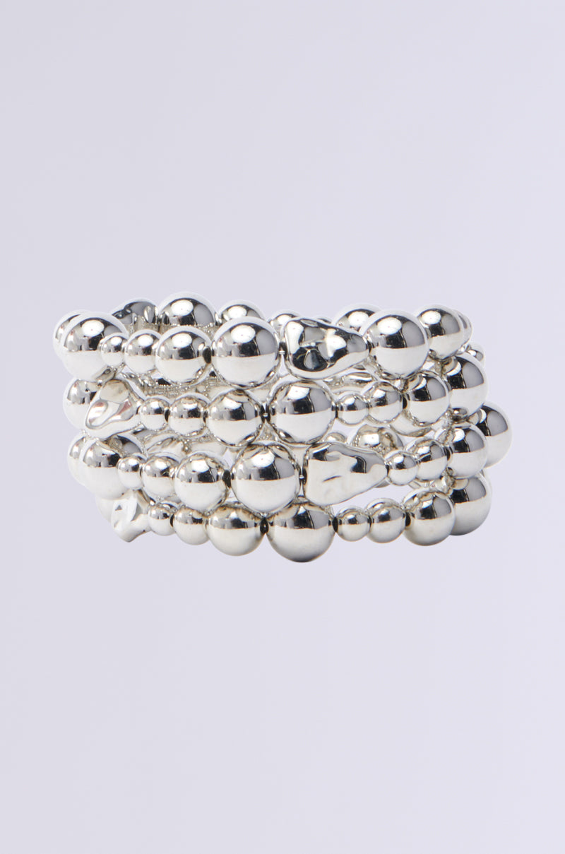 The BEADED GLORY BRACELET features a shiny silver ring coiled with metallic beads that reflect light, creating a lustrous appearance against a gray backdrop.