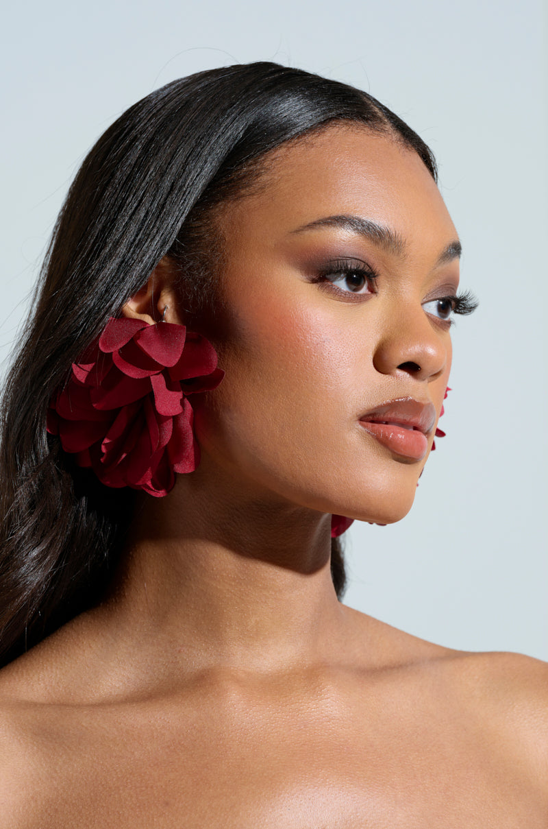 A woman with long dark hair models the Full Bloom Earrings, featuring oversized deep red floral silhouettes with a satiny finish. The earrings stand out against her neutral expression and the plain background in this shoulder-up view. 2