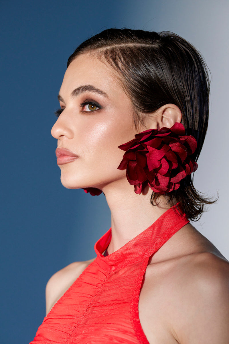A woman with short brown hair poses in profile against a blue and white gradient, wearing oversized FULL BLOOM EARRINGs and a bright red satin halter top. 0