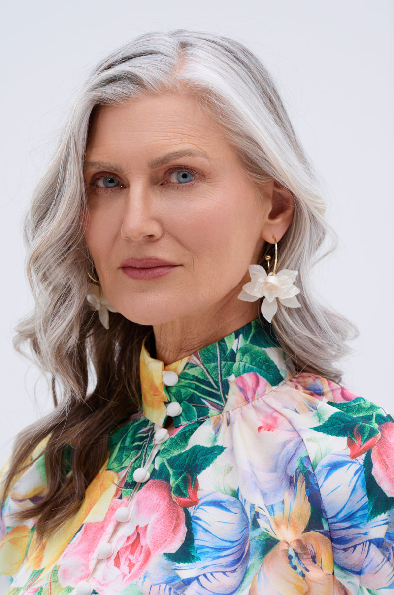 A woman with long gray hair and blue eyes wears a floral dress and WHITE FLORAL HOOP EARRING with pearlescent petals. She stands against a light background, her neutral expression embodying true botanical beauty style.