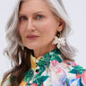 A woman with long gray hair and blue eyes wears a floral dress and WHITE FLORAL HOOP EARRING with pearlescent petals. She stands against a light background, her neutral expression embodying true botanical beauty style.