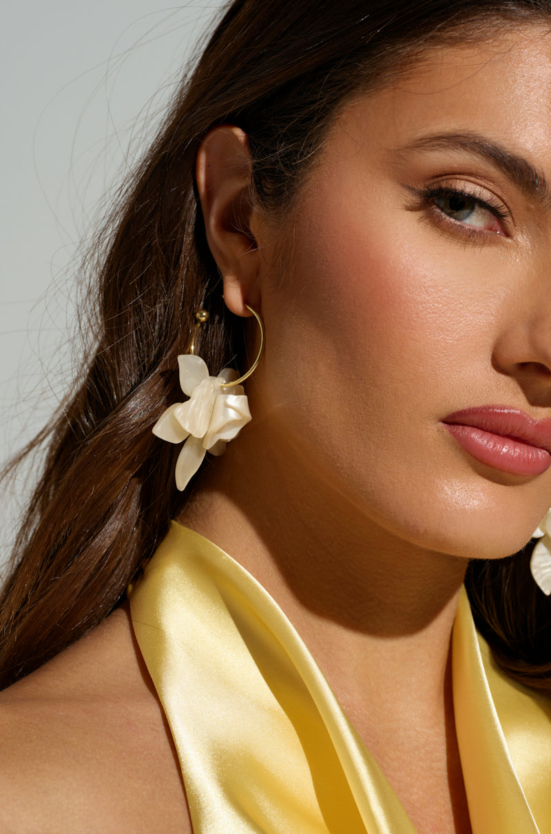A close-up shows a woman wearing the WHITE FLORAL HOOP EARRING, its pearlescent petals shimmering. Her long brown hair flows over a silky yellow top, all set against a plain light background that highlights the earring and her soft expression.