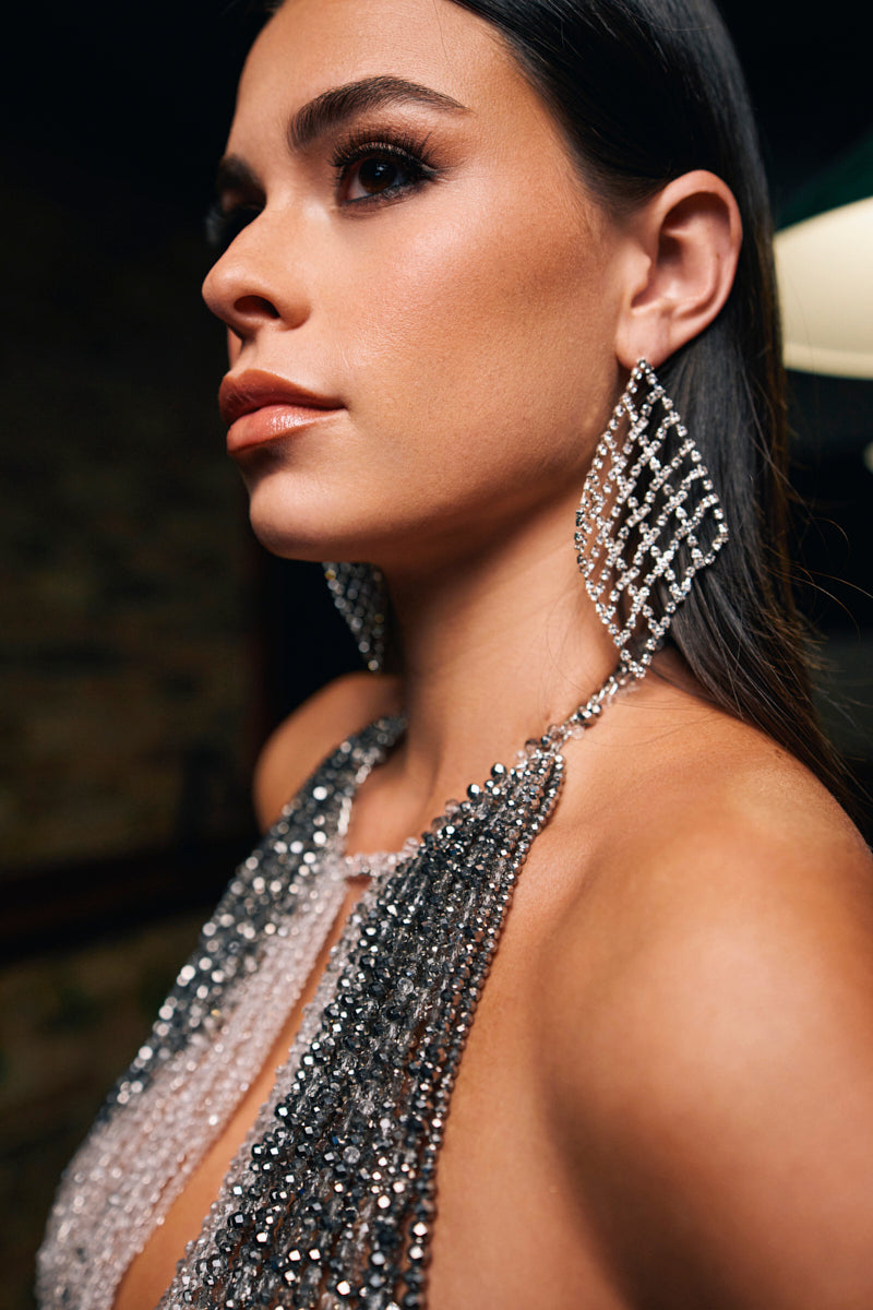 A woman with long dark hair and dramatic makeup wears the DANCE THE NIGHT AWAY EARRING—large, silver rhinestone earrings—along with a glittering beaded halter top, gazing confidently to the side against a softly lit background.