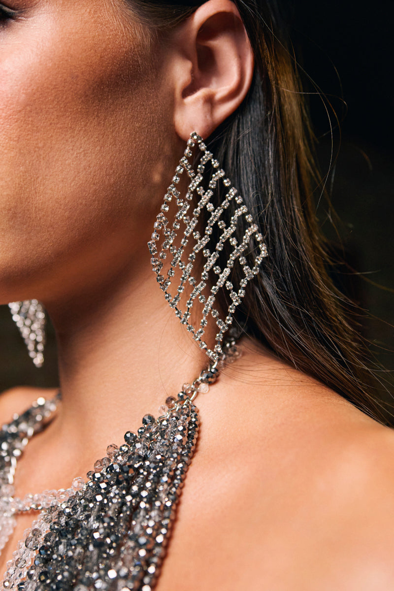 Close-up of a person wearing the DANCE THE NIGHT AWAY EARRING—large, dazzling silver rhinestone earrings. Their brown hair is tucked behind their ear, highlighting the statement accessory and a sparkly beaded garment.