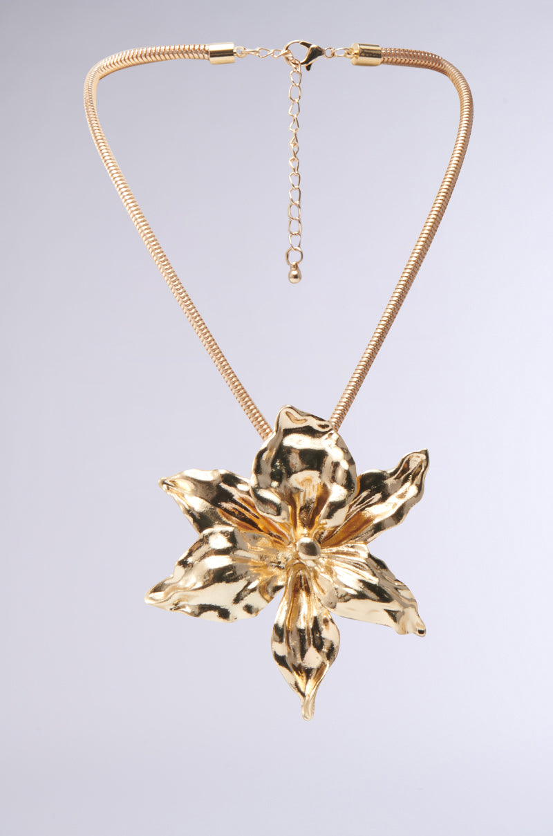 The IN THE SUN GOLD NECKLACE features a bold, detailed hibiscus-inspired pendant and is displayed against a simple light background.