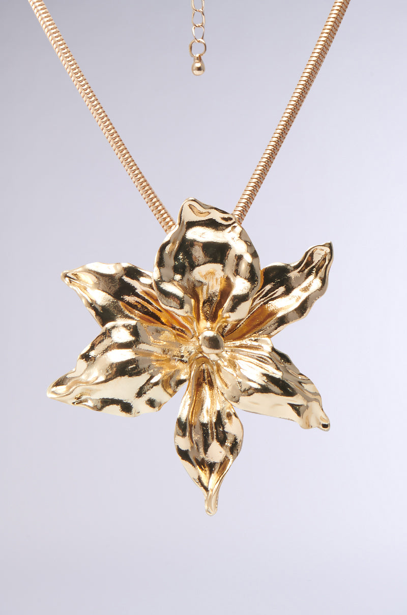 A close-up of the IN THE SUN GOLD NECKLACE, featuring a snake-chain and a large, textured five-petal hibiscus-inspired pendant, displayed against a light gray background.