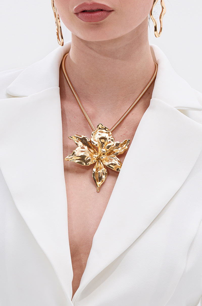 A woman wearing a white blazer with a deep neckline, gold hoop earrings, and the IN THE SUN GOLD NECKLACE featuring a large hibiscus-inspired pendant on a snake-chain.