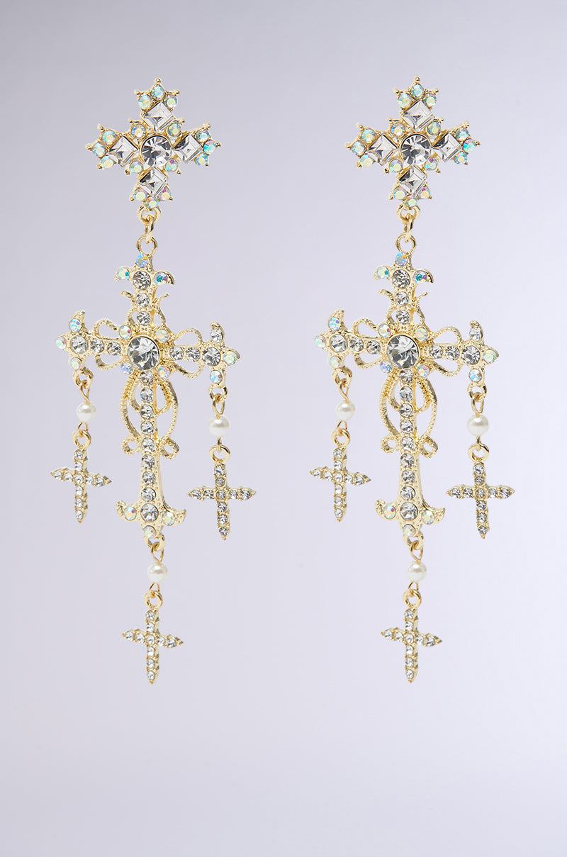 The DON'T BE CROSS EARRING features vintage-inspired ornate gold and silver with intricate cross designs, dangling floral motifs, and shimmering iridescent jewels set against a plain gray background.