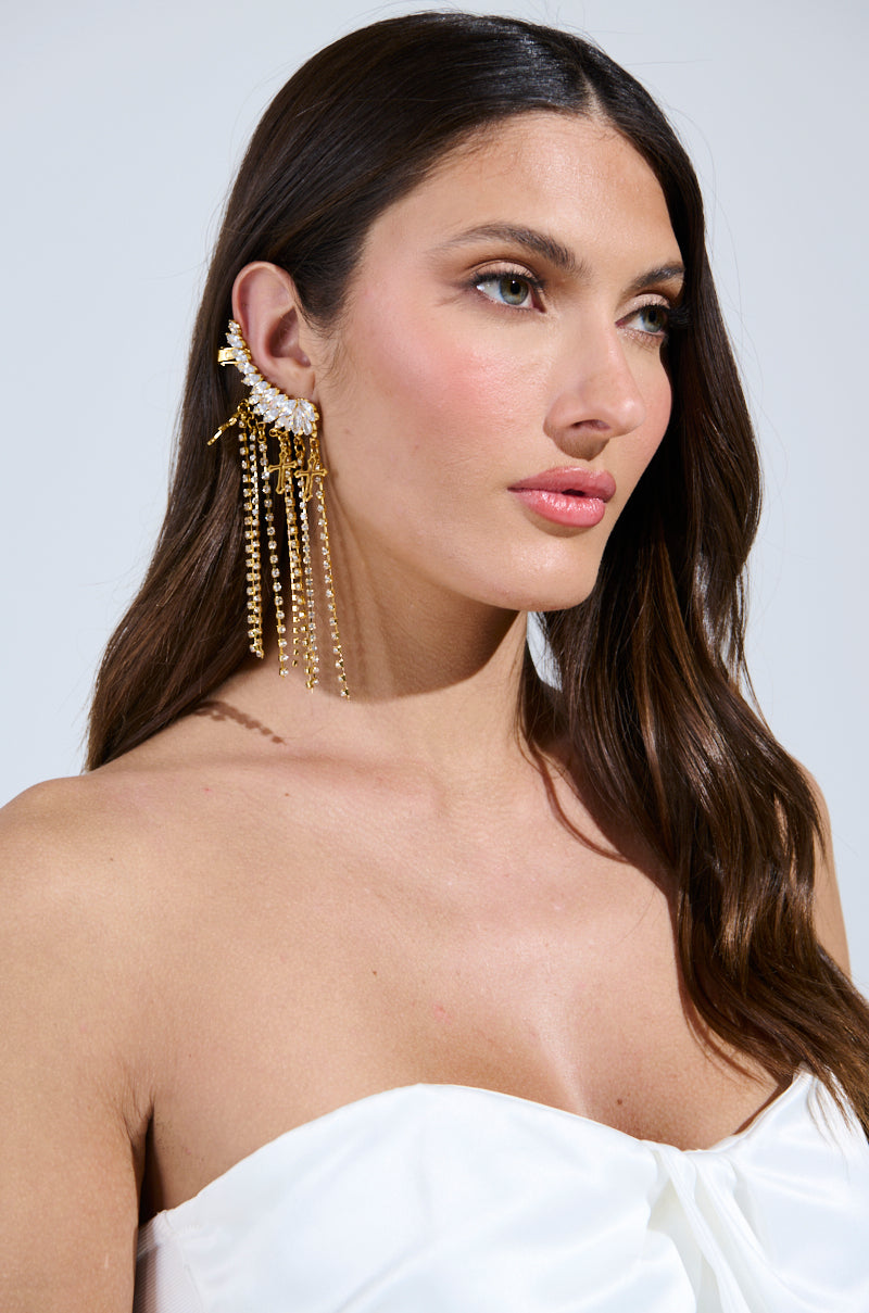 A woman with long brown hair, wearing a strapless white top, dons the ANGEL VIBES RHINESTONE CUFF on her left ear. Adorned with cascading chains and gold cross charms, she exudes angelic vibes through her calm expression while gazing slightly to the side.