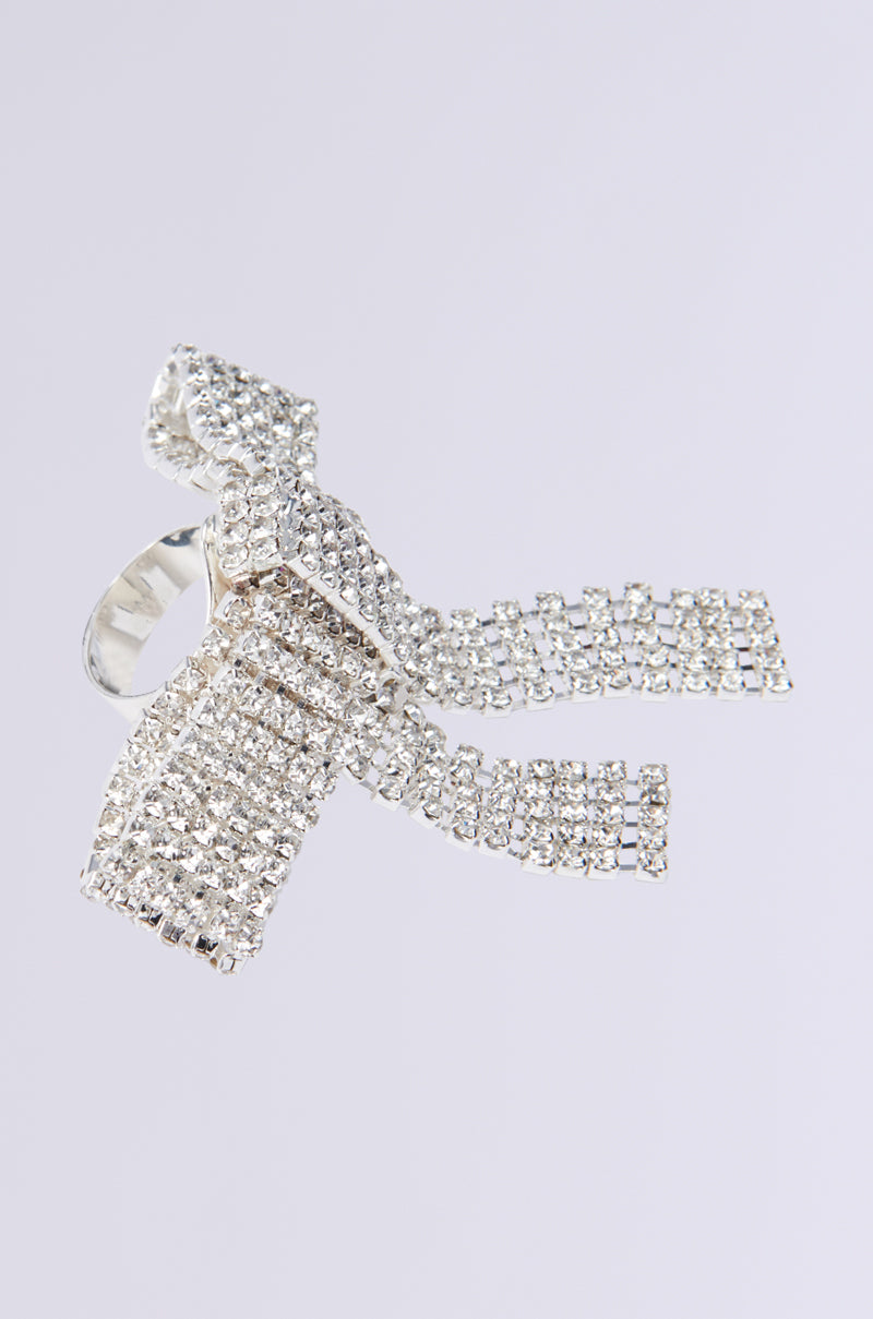 The TIE WITH A BOW STATEMENT RING is a silver piece adorned with small, sparkling rhinestones in an intricate bow design. Its long draped tails and adjustable band present an elegant and luxurious look against a plain white backdrop.