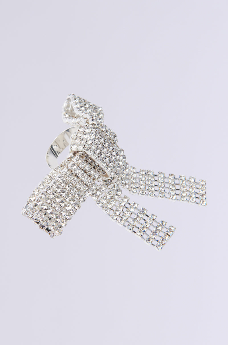 The TIE WITH A BOW STATEMENT RING showcases a striking silver bow adorned with sparkling rhinestones. Its central knot and flowing tails sit on an adjustable band for the perfect fit, set against a plain light gray backdrop.