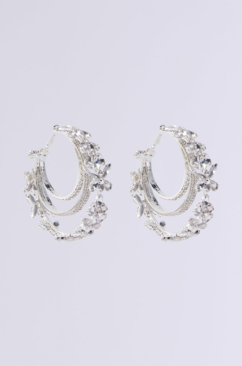 The DRAMA IS MY MIDDLE NAME HOOP earrings are silver hoops adorned with intricate rhinestones, featuring layered and sparkling designs for an elegant, luxurious look against a plain gray backdrop.
