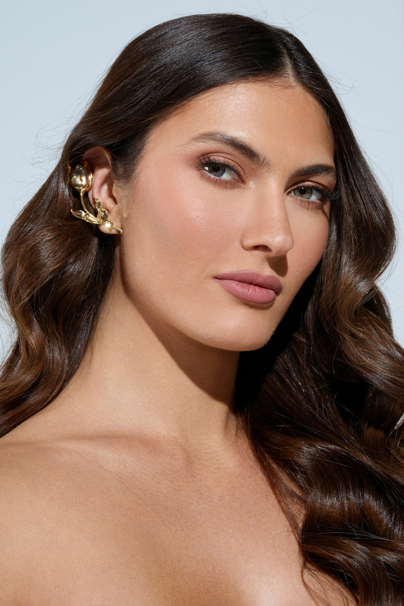 A woman with long, wavy brown hair wears an elegant ROSEY EARRING in faux gold. She gazes softly at the camera, highlighting her smooth makeup and subtle lipstick. The neutral background accentuates her features and accessory. 0