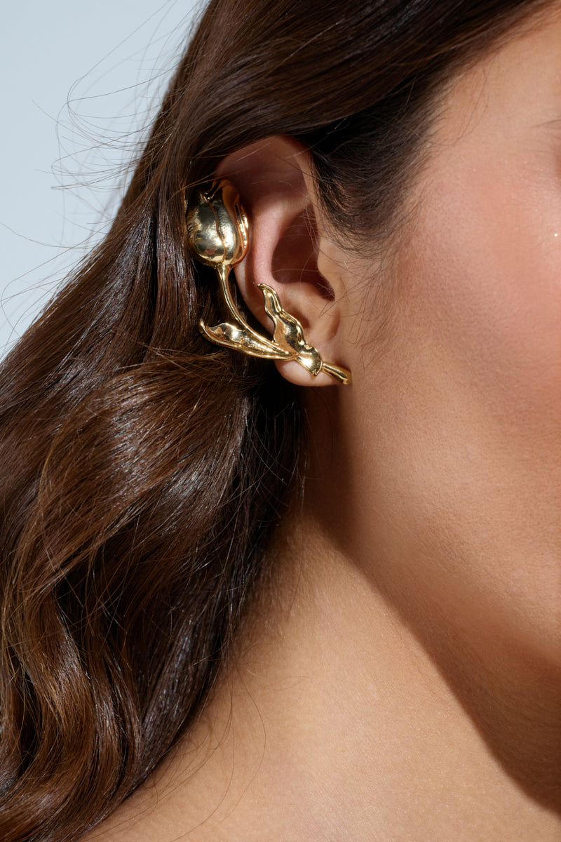 A close-up image shows a woman's ear adorned with the ROSEY EARRING, a faux gold ear cuff in a rose-flower design, featuring a pearl-like detail and an elegant ear-climbing silhouette. Her brown hair cascades down the side. 1