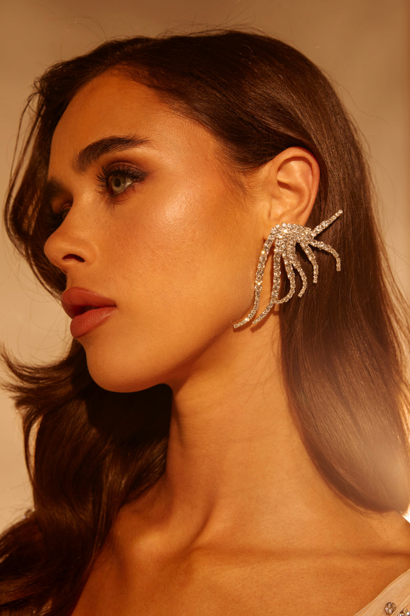 A woman with wavy brown hair wears the IN MY ZONE SILVER EARRING, featuring large, sparkling rhinestone strands shaped like trailing tendrils. She has smooth skin and natural makeup, shown in warm lighting. 0