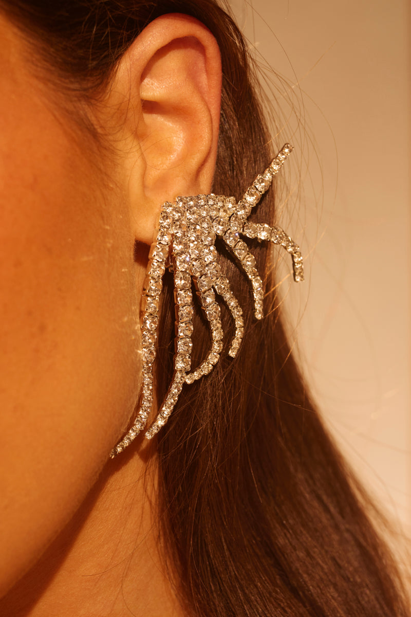 Close-up of a person's ear adorned with the IN MY ZONE SILVER EARRING: a large, sparkling silver accessory with abstract branching spikes encrusted with rhinestones. Brown hair is neatly tucked behind the ear. 1