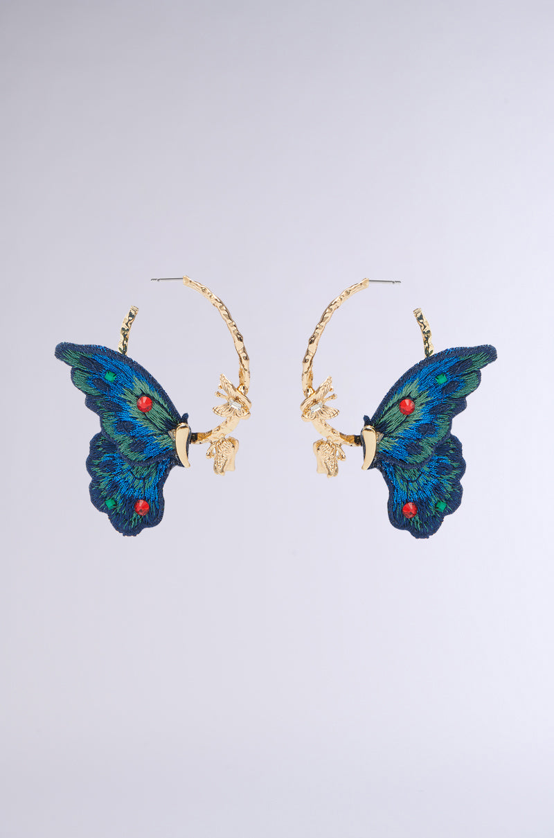 The SO FLY BUTTERFLY HOOP EARRING features vibrant blue and green embroidered textile wings with gold-tone elements and rhinestone accents, set symmetrically on a light gray background.