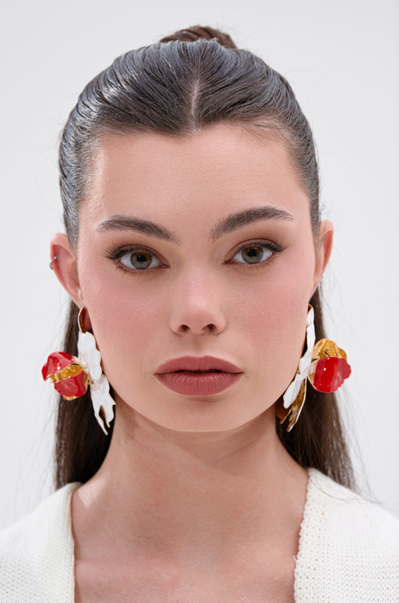 A woman with straight brown hair pulled back, wearing the bold red and white FLORAL GARDEN EARRING with an oversized floral motif and a white top, looks directly at the camera against a plain light background. 2