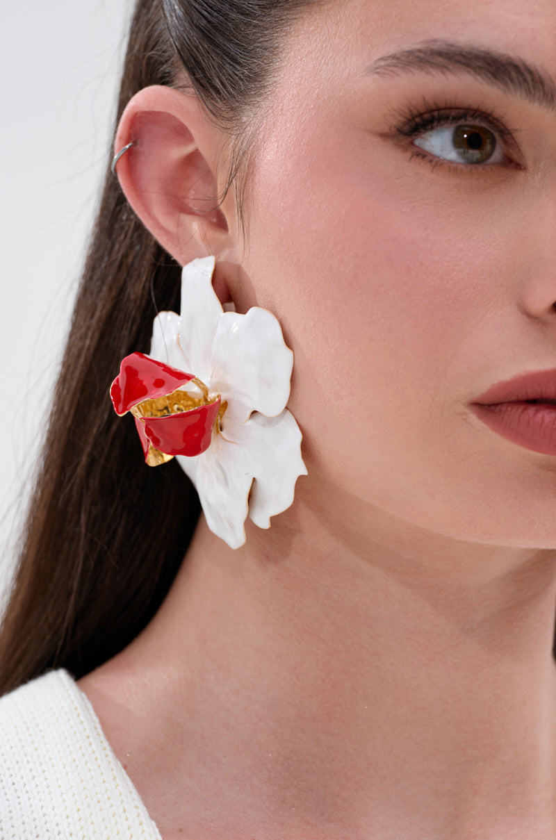 A close-up of a woman in a white top and pulled-back brown hair wearing the FLORAL GARDEN EARRING, featuring an oversized floral motif with sculptural textured petals and a red and gold center. 0