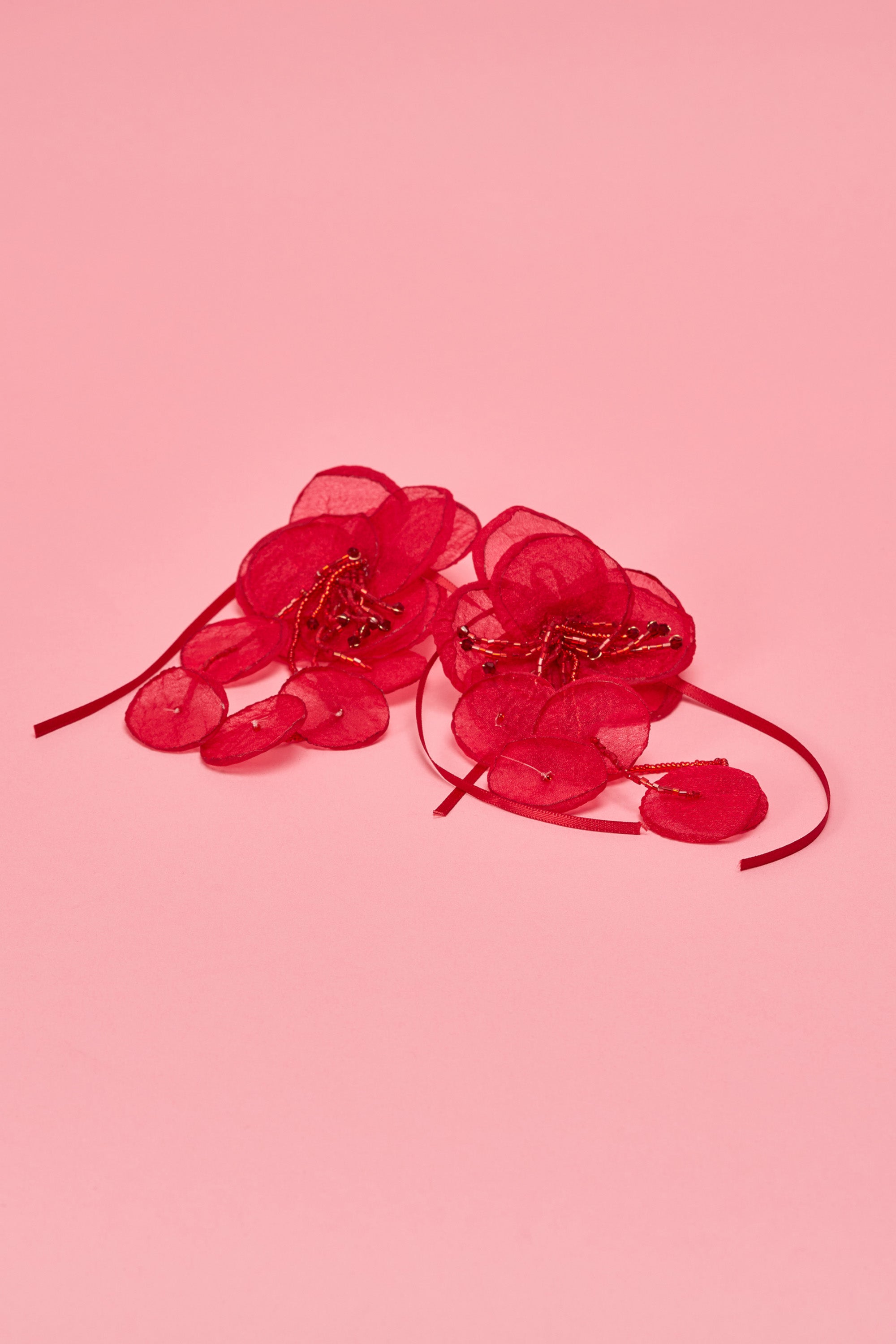 The SIPPIN WINE EARRING features two red sheer organza petals arranged as floral earrings with ribbon and beaded centers, set against a pink background. 0