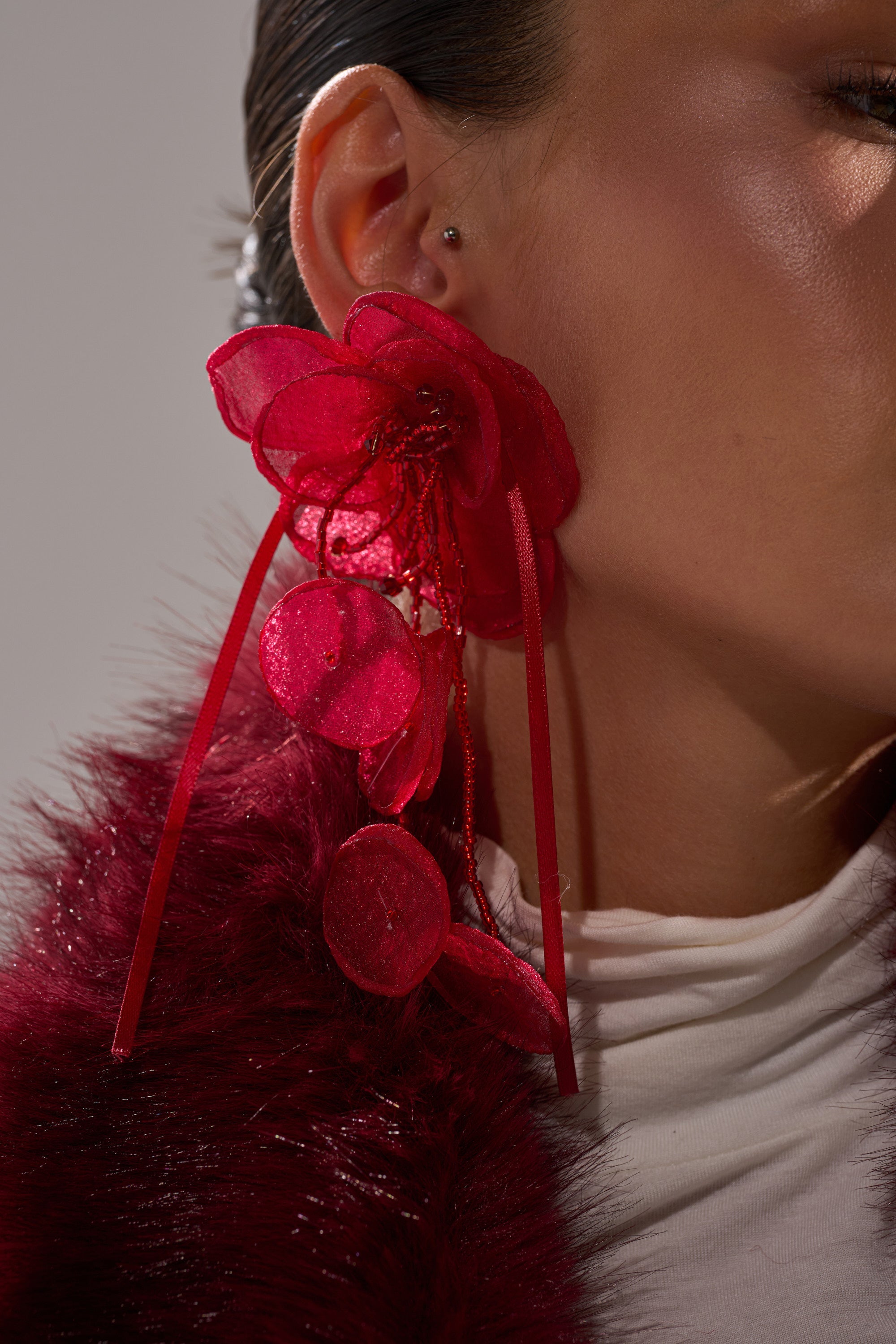 Close-up of a person wearing the bold red SIPPIN WINE EARRING. Smooth skin, slicked-back dark hair, a burgundy faux fur garment, and a white turtleneck complete the stylish look. 2