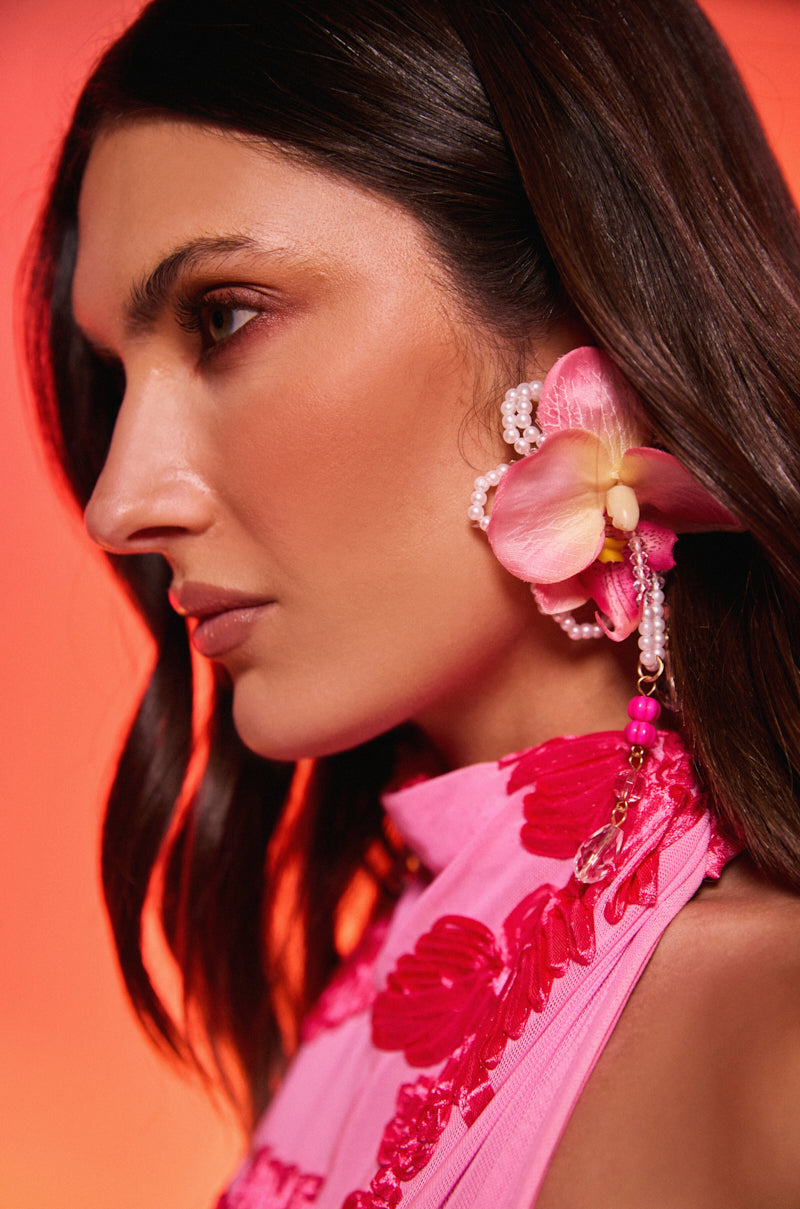 A woman with long dark hair wears a pink floral top and the bold TO THE TROPICS EARRING, adorned with beads and floral accents. Photographed from the side against a warm red background, this look is perfect for resort wear inspiration. 0