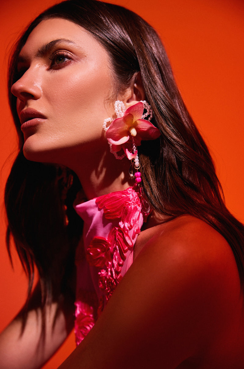A woman with long brown hair poses against an orange background, wearing a pink floral top and the TO THE TROPICS EARRING adorned with pink and yellow flowers, her features highlighted in dramatic lighting for resort wear chic. 1