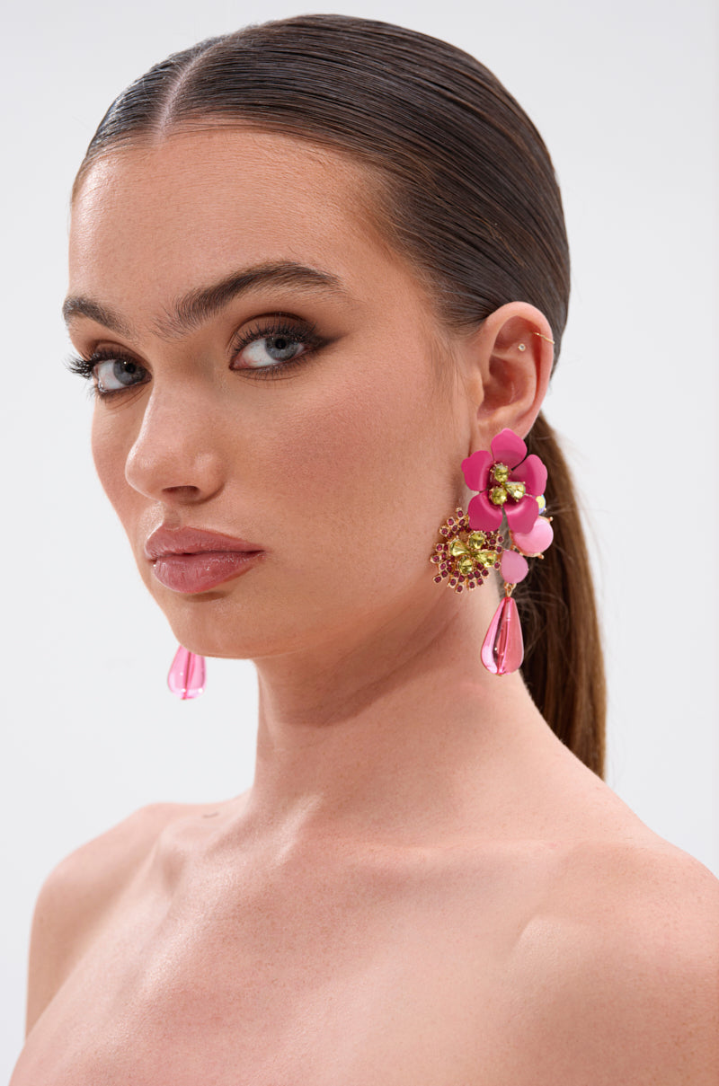 A woman with slicked-back brown hair wears bold pink BLISS EARRINGs—floral statement pieces with dangling teardrop beads and sparkling crystal centers—paired with natural makeup, defined brows, and winged eyeliner against a plain background. 1