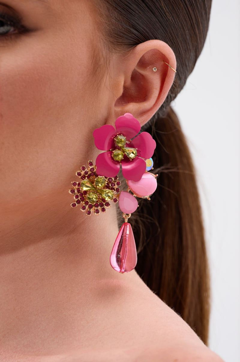 Close-up of a woman's ear wearing the BLISS EARRING, a floral statement piece with pink and yellow flowers, glittering crystal centers, and a dangling pink teardrop bead. Her hair is pulled back tight. 3