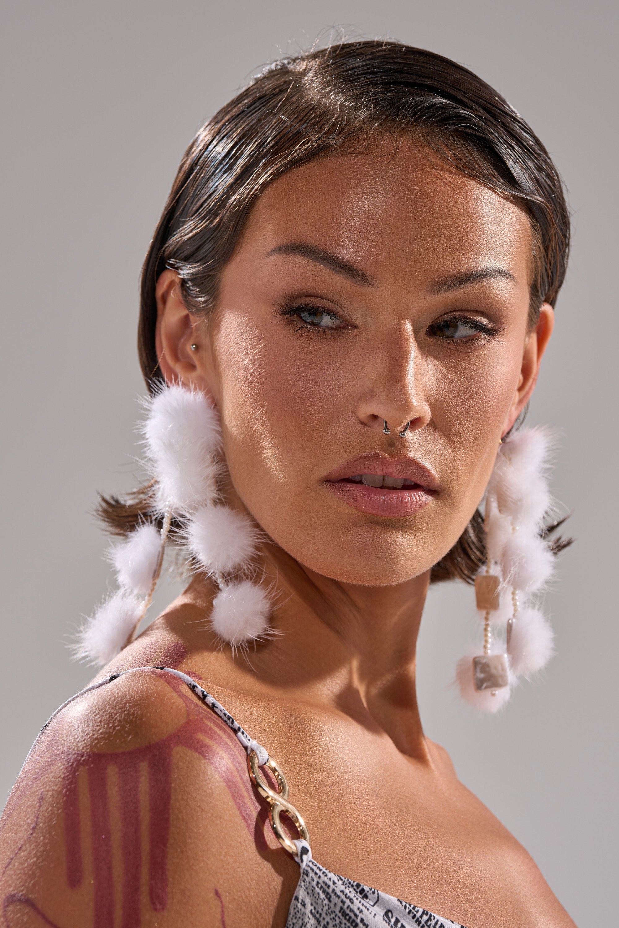 A woman with short dark hair wears SNOW PRINCESS EARRING—large white faux fur pom-poms—and a patterned thin-strapped top. She has a septum piercing and gazes to the side with a neutral expression against a plain background. 1
