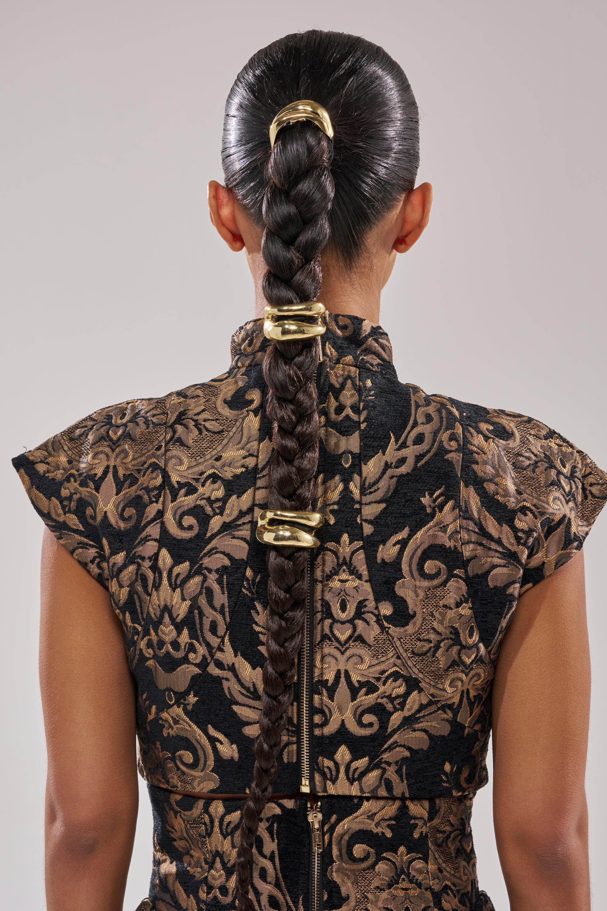 A person with long, dark hair in a sleek bubble ponytail styled with the GOLDEN RULE HAIR TIE SET, wearing a high-collared, short-sleeved garment featuring an ornate black and gold pattern, shown from the back.