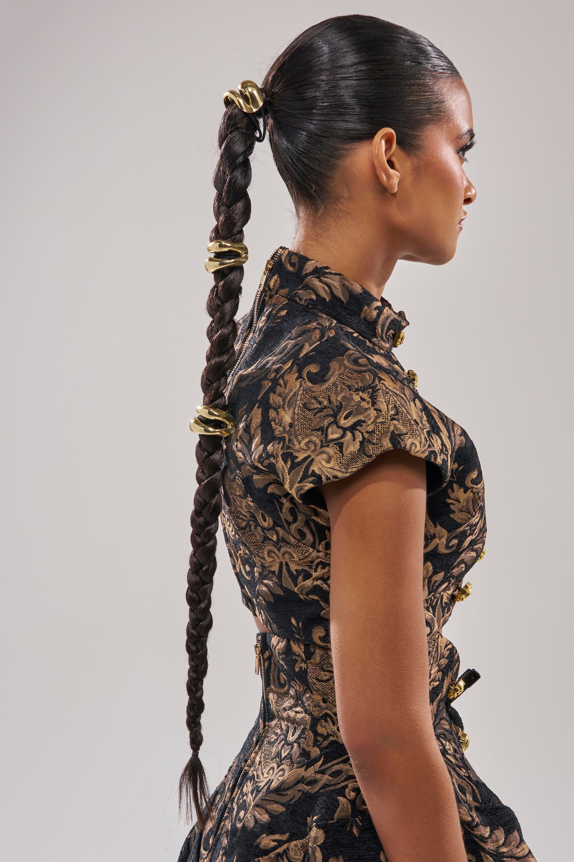 A woman with a long ponytail secured by the GOLDEN RULE HAIR TIE SET wears a short-sleeved black and gold brocade dress, standing in profile against a neutral background.
