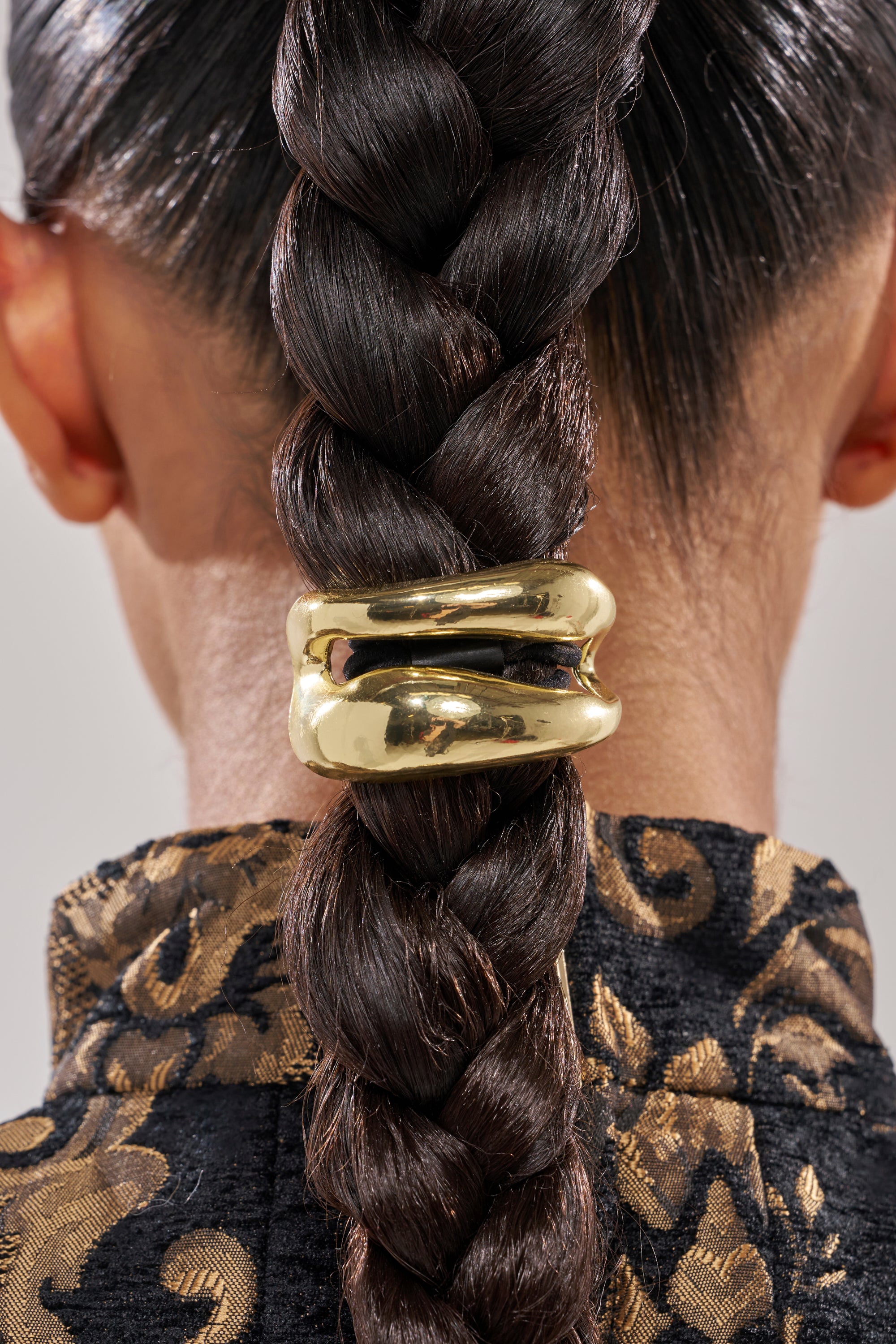 A person with dark braided hair secured by the GOLDEN RULE HAIR TIE SET. They wear a dark, textured garment with a gold floral pattern. The image captures the back of their head and neck.