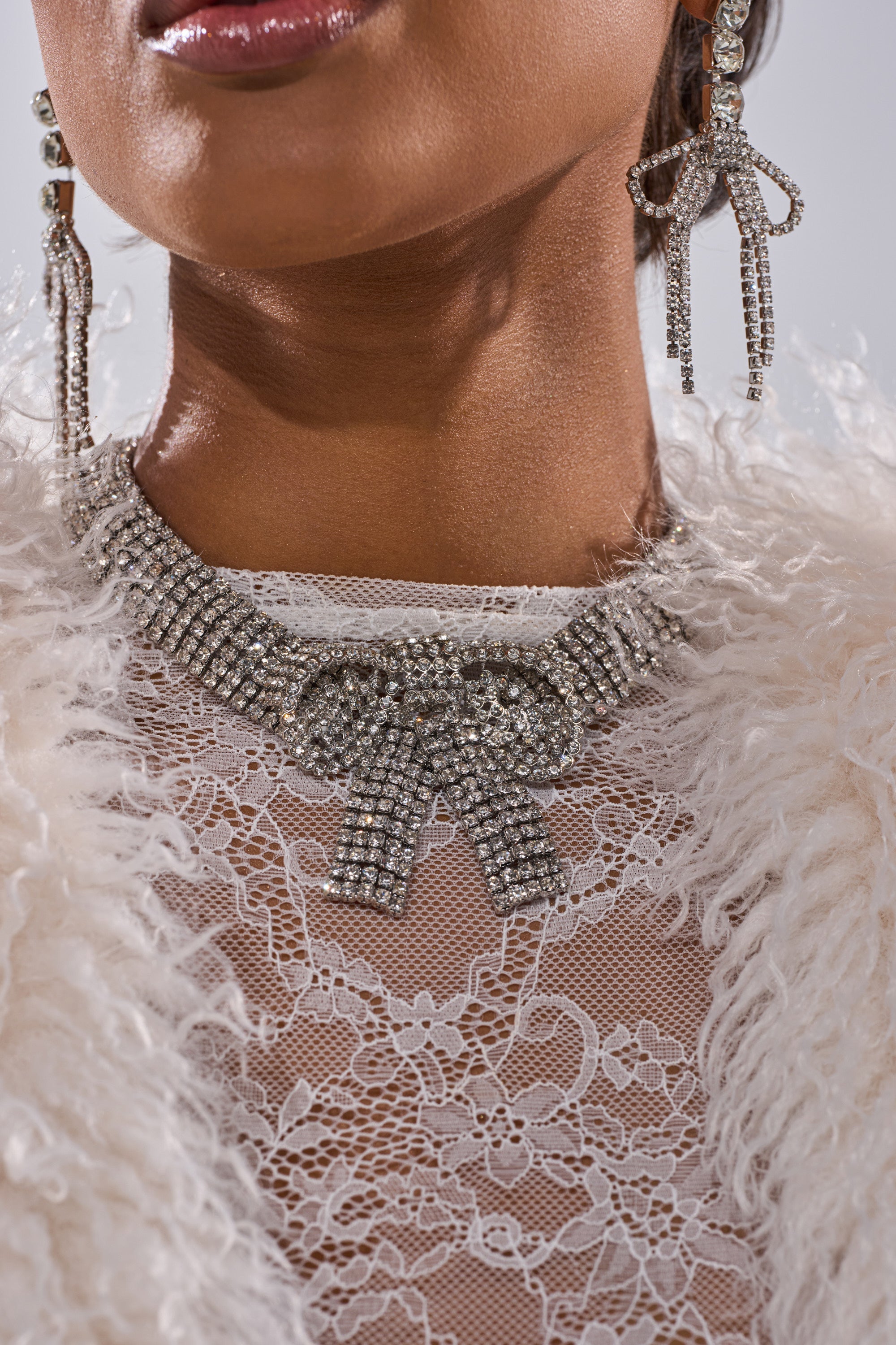 A close-up of a person wearing the GABBY BOW NECKLACE with matching rhinestone earrings. The statement necklace pairs elegantly with their white lace top and a textured, fluffy off-white garment. 1