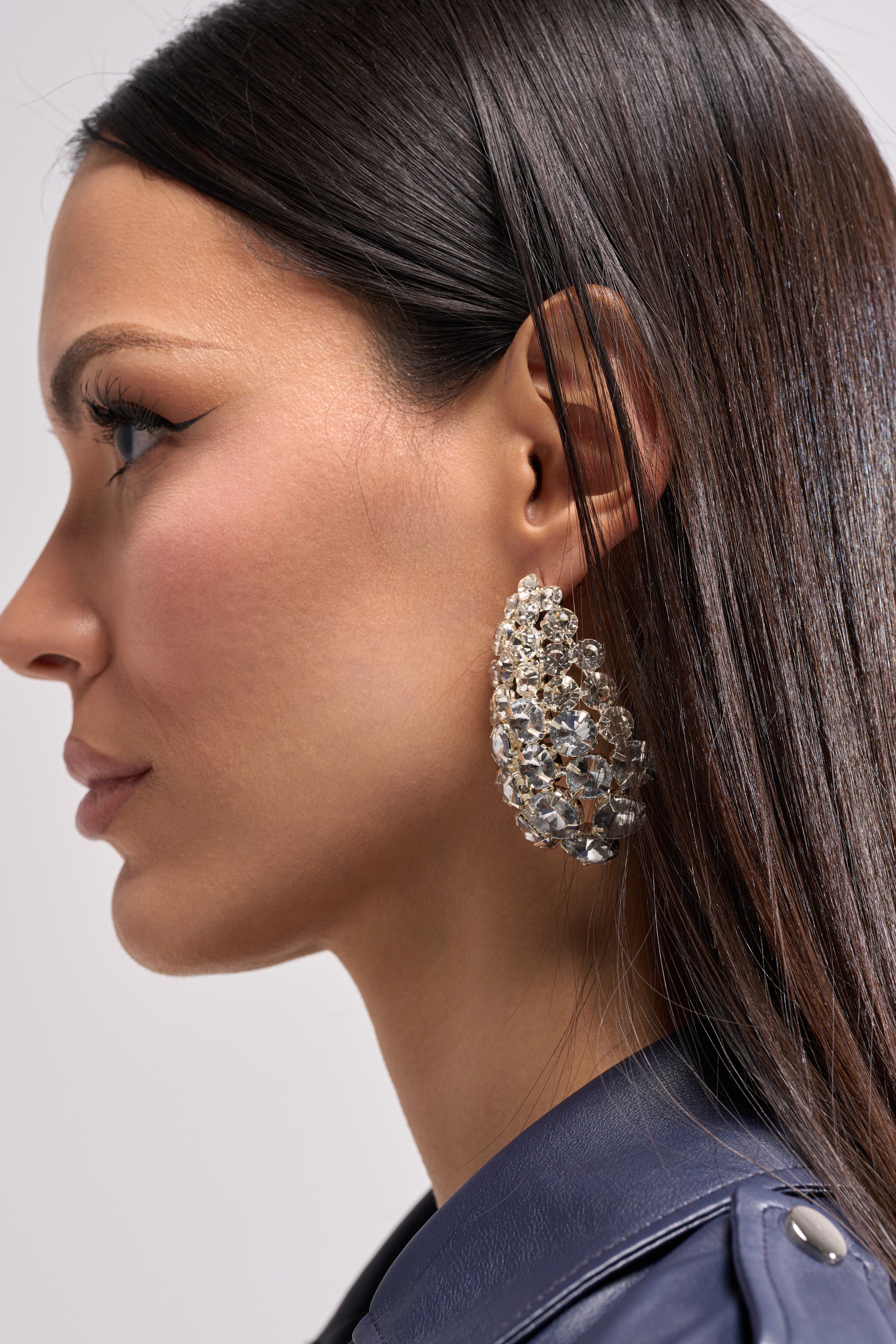 A woman with straight dark hair wears the NIGHT IS YOUNG EARRING—a sparkling, bold statement piece ideal for glamorous evenings—paired with a dark leather jacket, shown in profile against a light background.