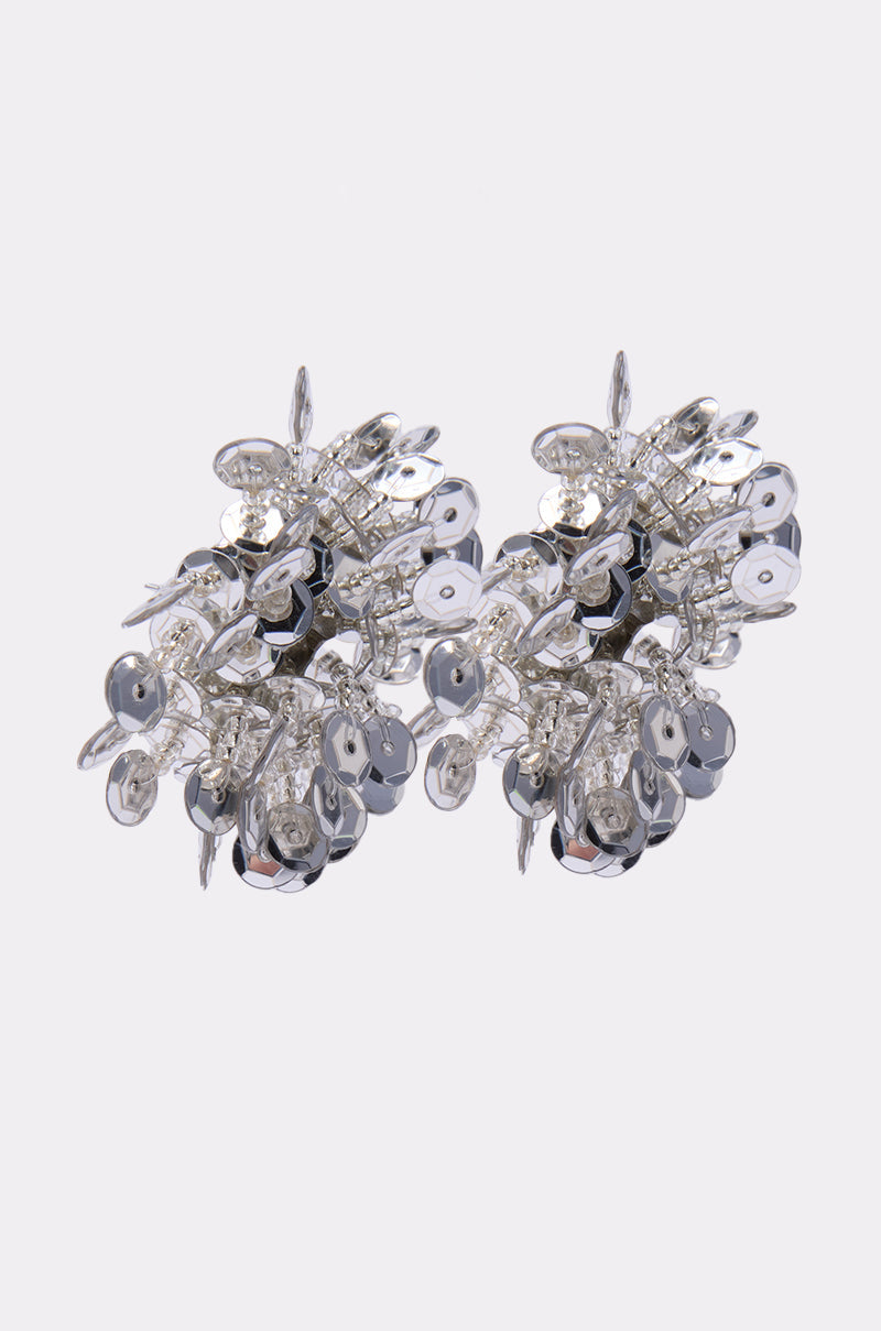The STELLAR EARRING is a pair of silver, cluster-style earrings featuring shiny sequins and silver-tone accents, creating a textured and reflective look against a plain white background. 2