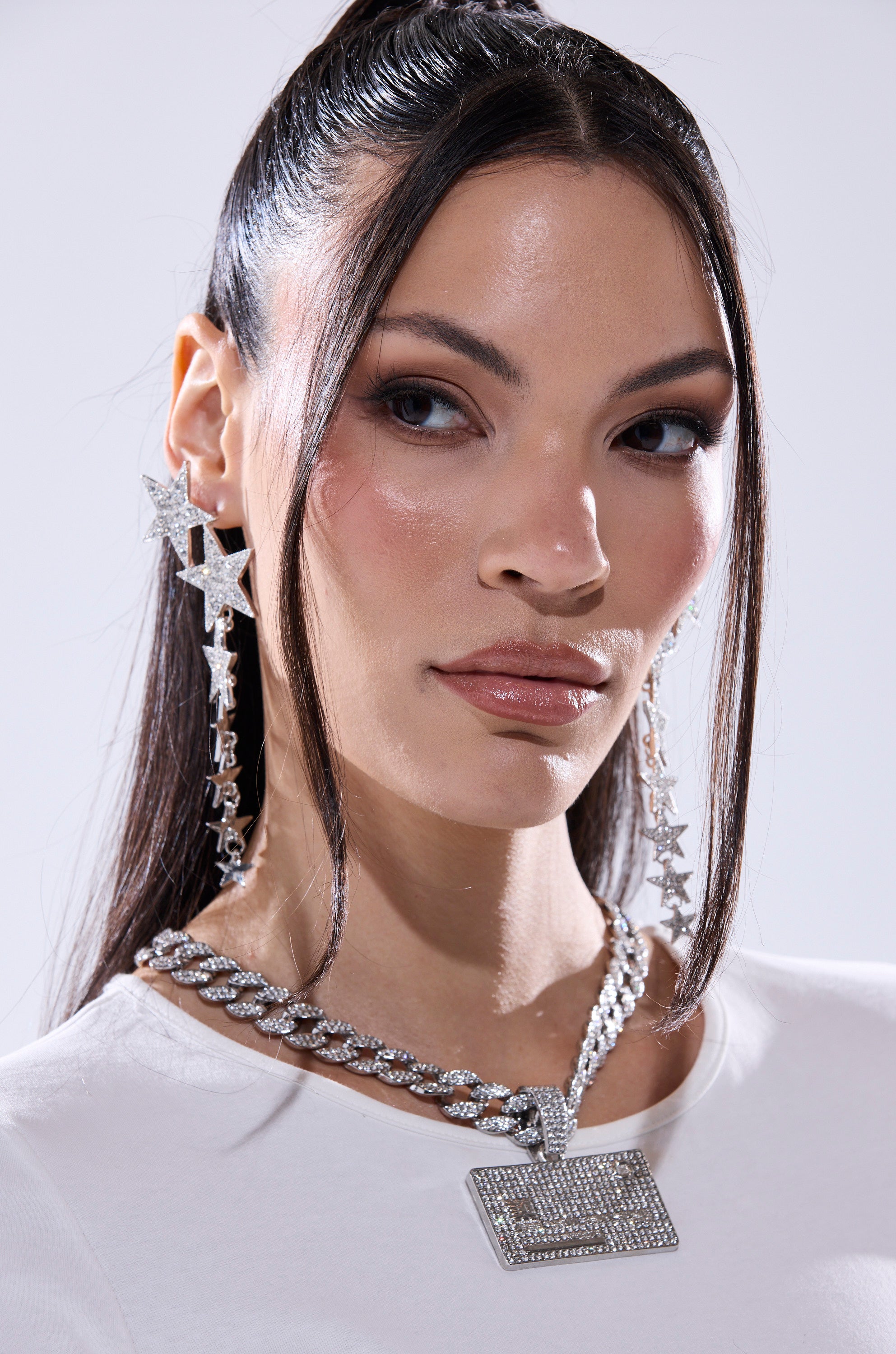 A woman with long, straight dark hair and smooth skin wears sparkly STARGURL EARRING with rhinestone accents and two silver chain necklaces with rectangular pendants. Dressed in white, she looks confidently at the camera.