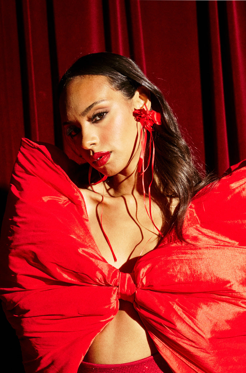 A person poses confidently before deep red curtains, wearing a dramatic red bow top and bold lipstick. Their long, dark hair is styled with LACED UP EARRING satin ribbon earrings for a playful, elegant touch.