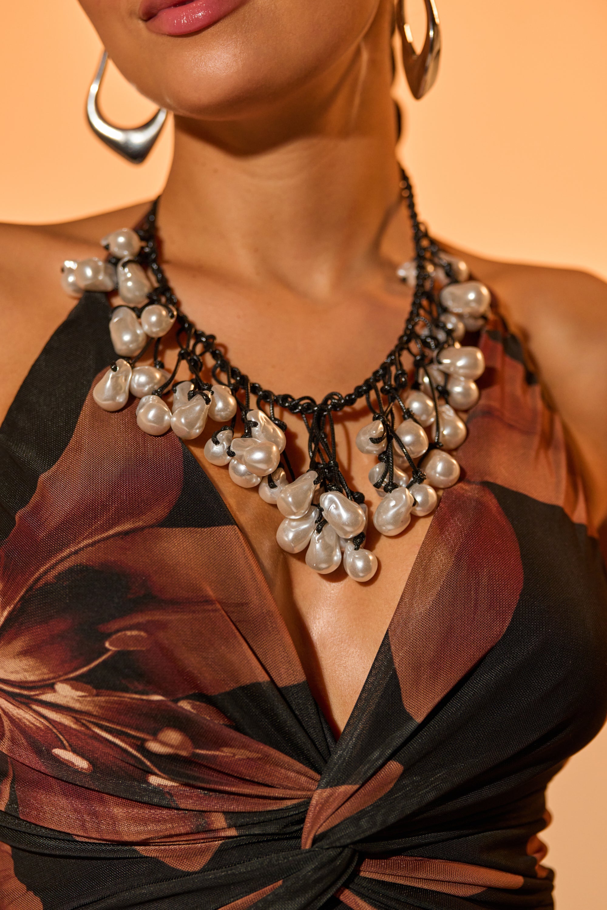 A woman wears a floral dress with a deep neckline and the GIVING EVERYTHING NECKLACE, featuring large faux pearls and a black chain, against a warm beige background. She completes the look with silver hoop earrings.