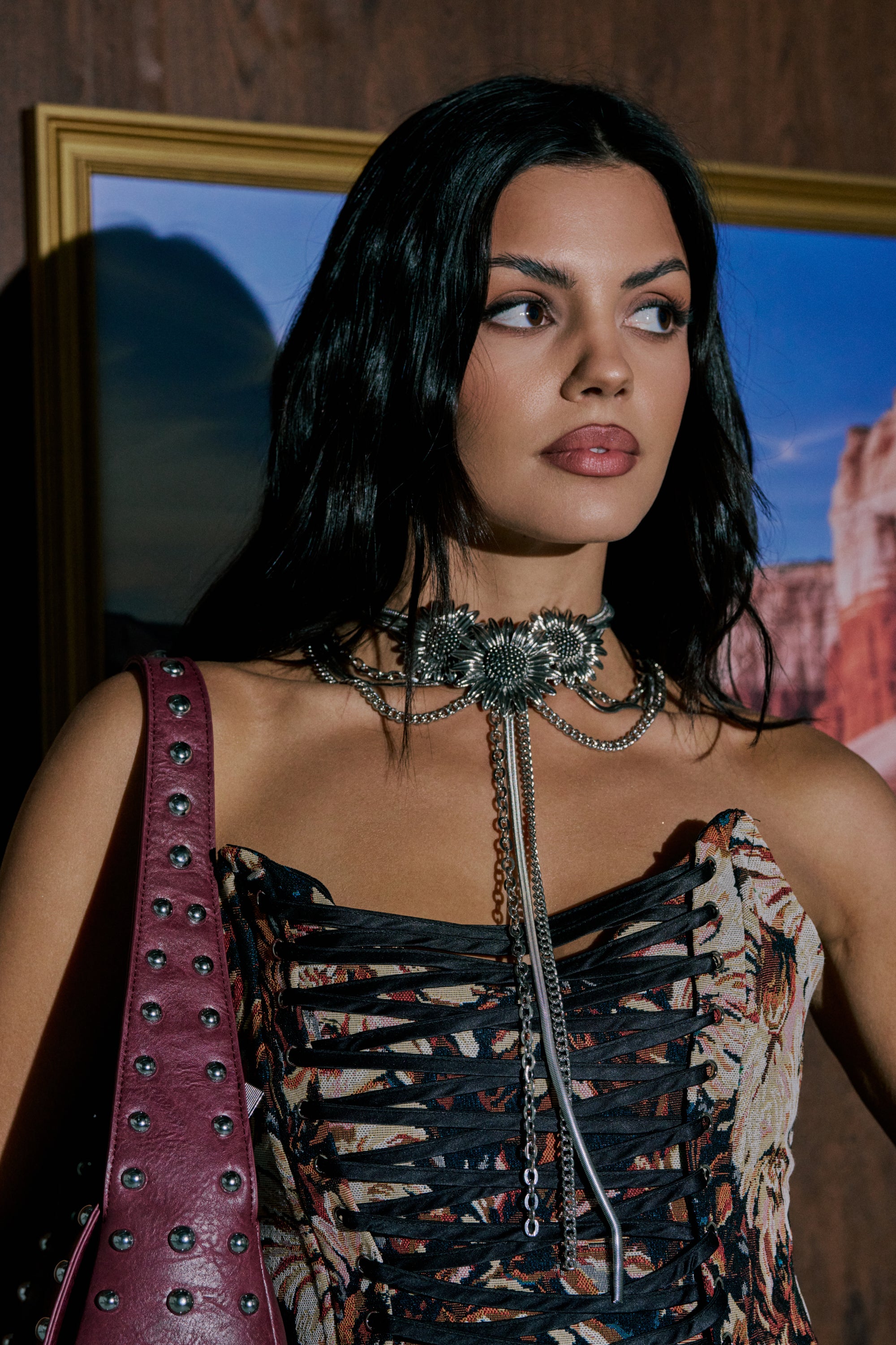 A woman with long dark hair wears a patterned, corset-style top and the NOT KANSAS ANYMORE NECKLACE. She holds a maroon studded bag and stands in front of a framed landscape photo on a wooden wall. 0