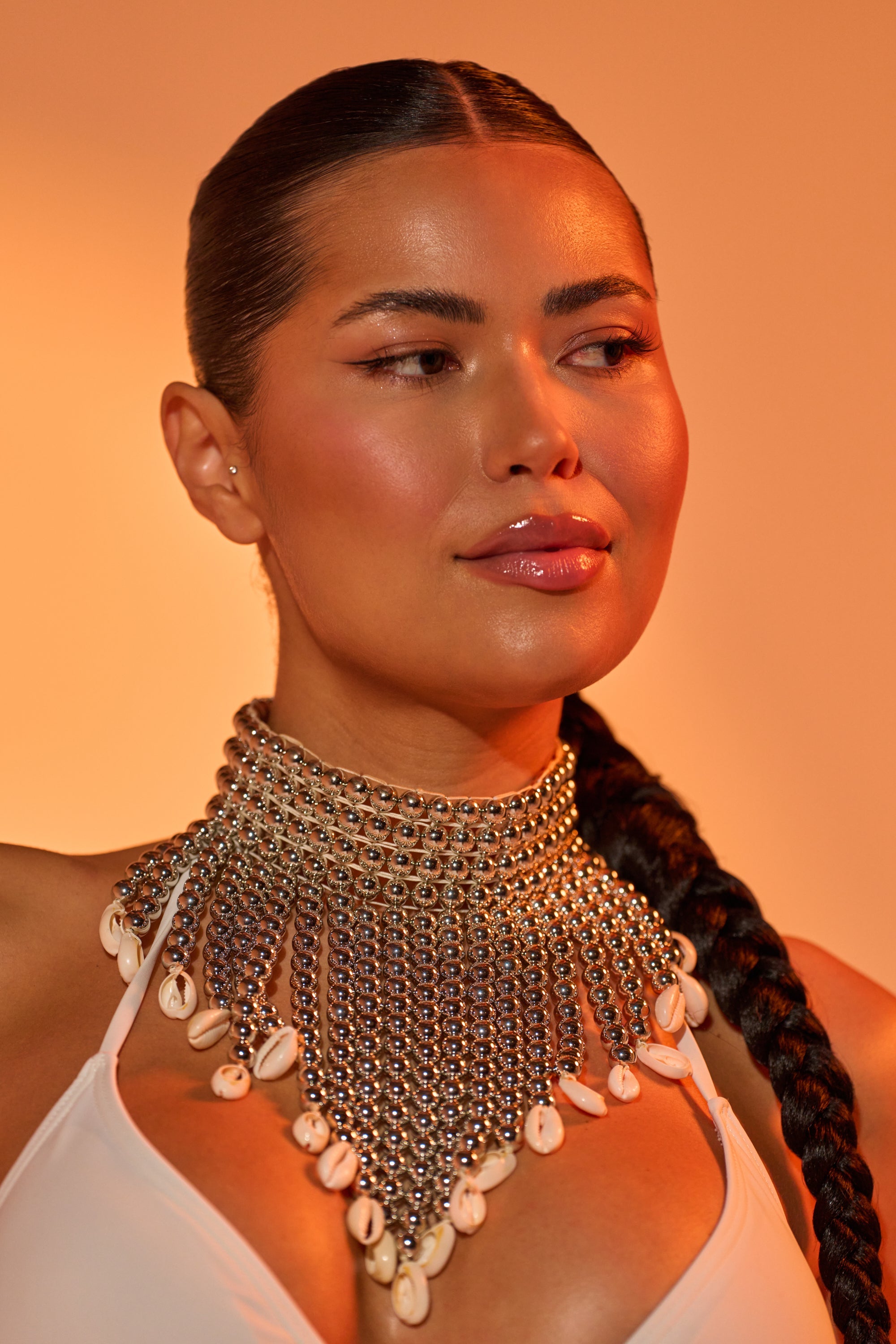 A woman with glowing skin and long braided hair wears a white top and the TOO CHIC SEASHELL NECKLACE—an eye-catching piece made of silver beads and white shells—set against a warm orange background. 0