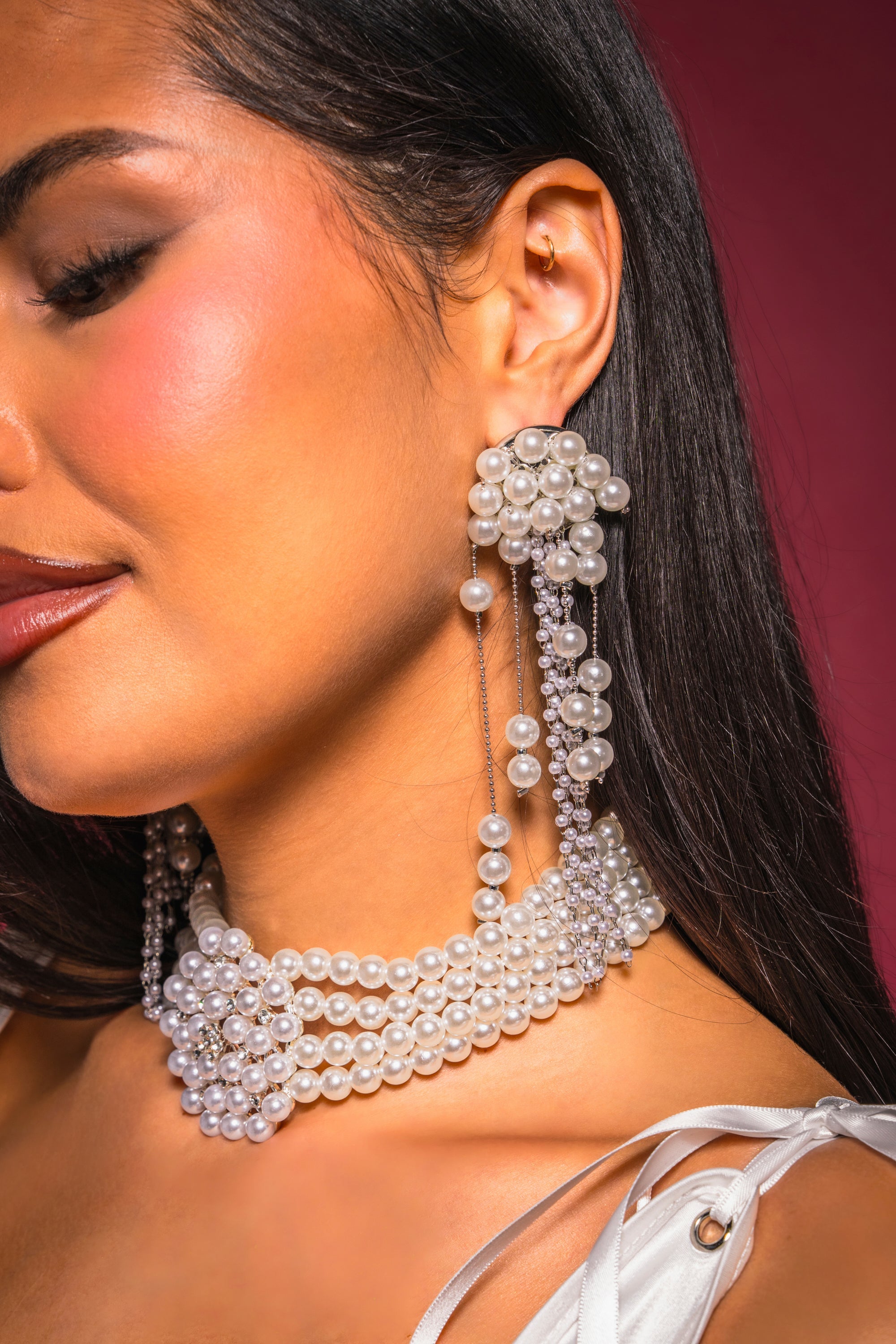 Close-up of a woman wearing the DRIPPIN IN PEARLS EARRING, featuring statement pearl tassels. She pairs it with a matching pearl choker and white thin-strap top, against a deep pink background. 0