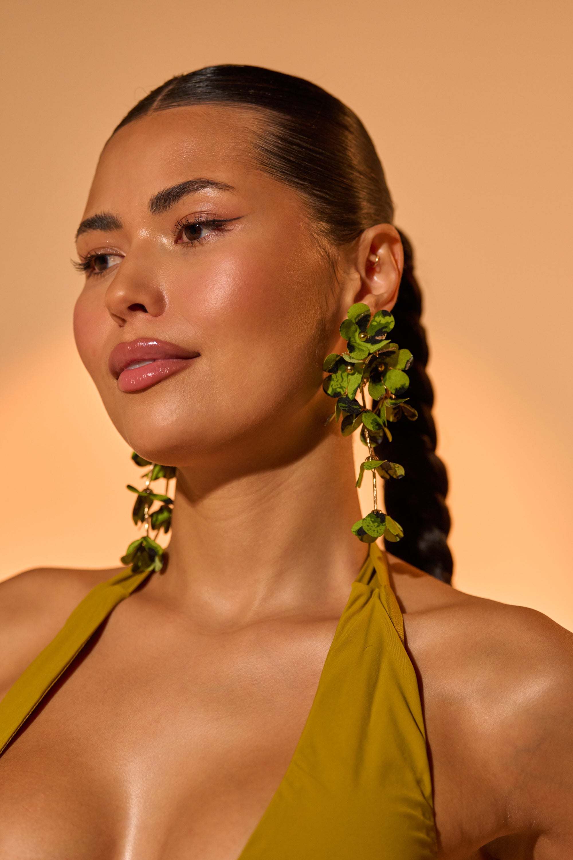 A woman with smooth, glowing skin and sleek, braided hair wears the bold CATCH ME IF U CAN EARRING and an olive green halter top, gazing to the side against a warm beige background. 0