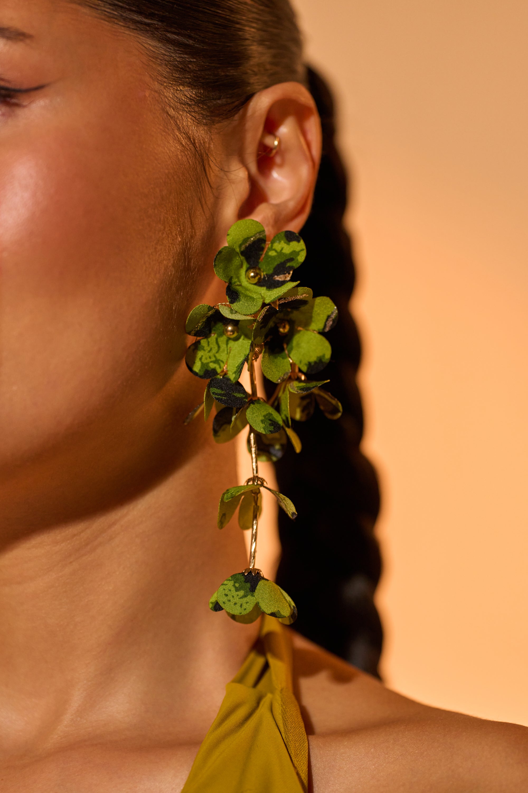 Close-up of a person wearing the CATCH ME IF U CAN EARRING—bold green leaf earrings with gold accents. Their braided dark hair and yellow top stand out against a warm, neutral background, emphasizing these statement earrings. 1