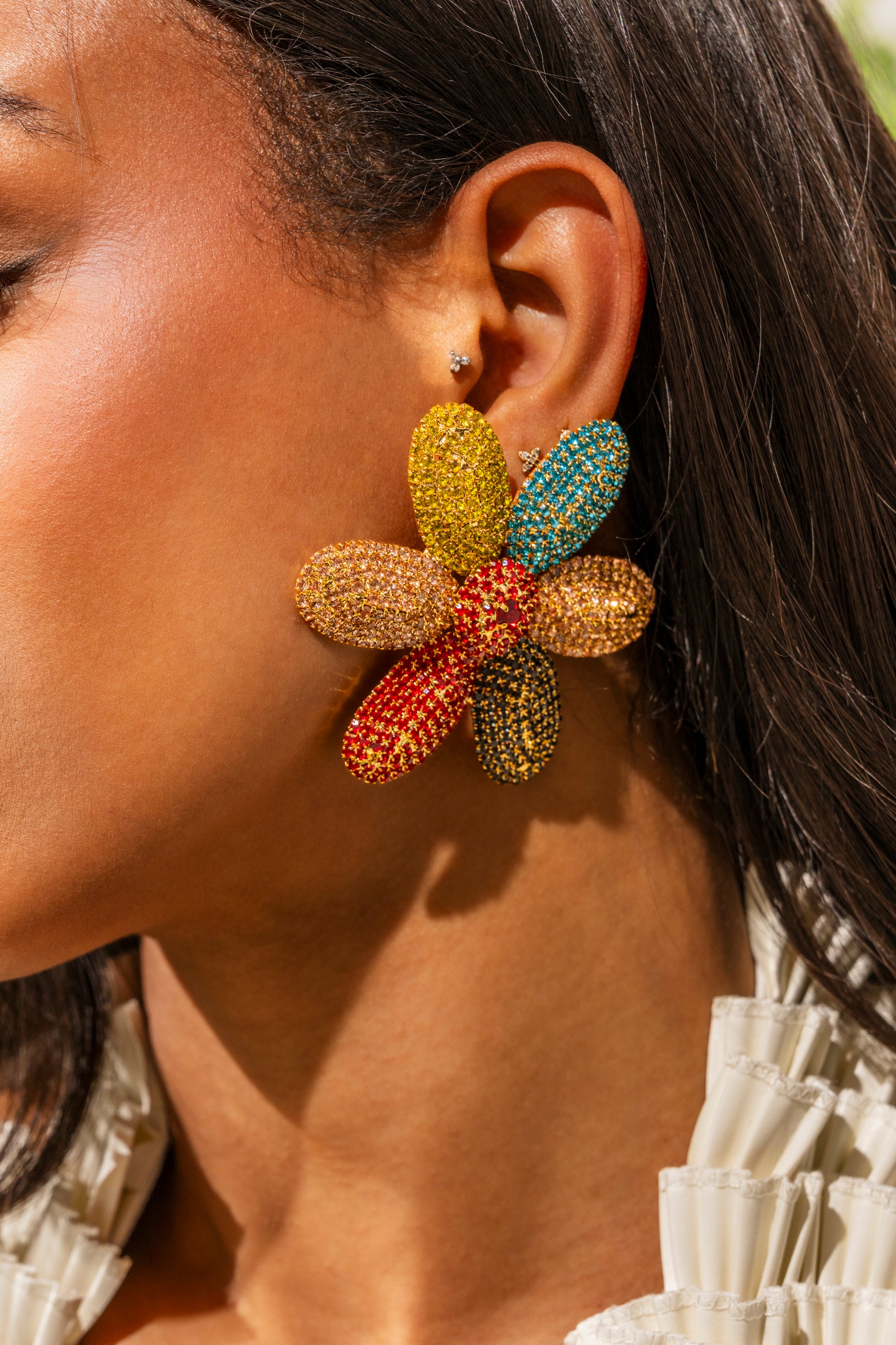 A close-up of a woman's ear displays the TAKE ME TO JAPAN EARRING—large, multicolored statement flowers with jeweled yellow, red, blue, and orange petals—paired with a small stud above for extra sparkle. 0