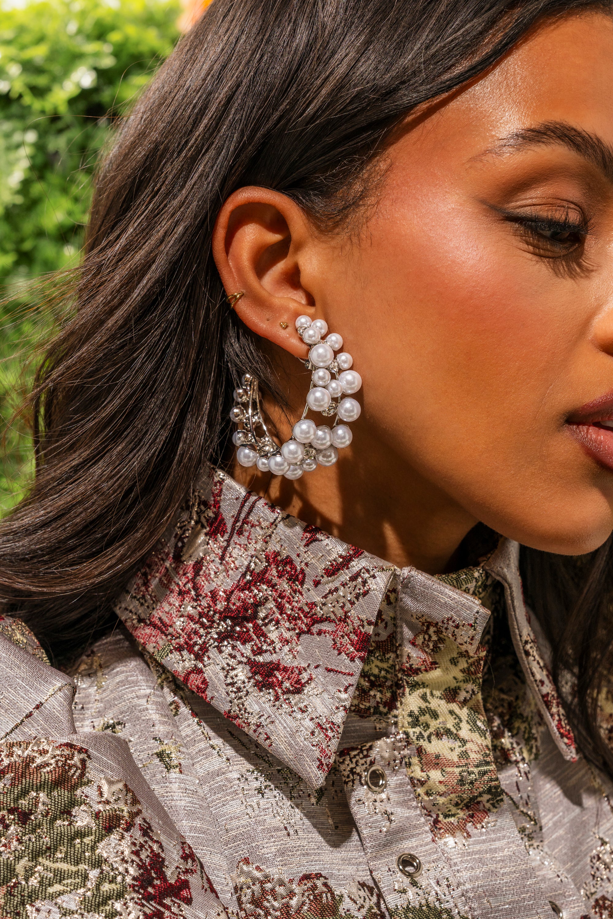 Close-up of a woman wearing the PEARL PLZ EARRING, paired with small gold studs and a patterned collared shirt. Her dark, wavy hair frames her face as she stands outdoors among greenery, showcasing chic bridal style. 0