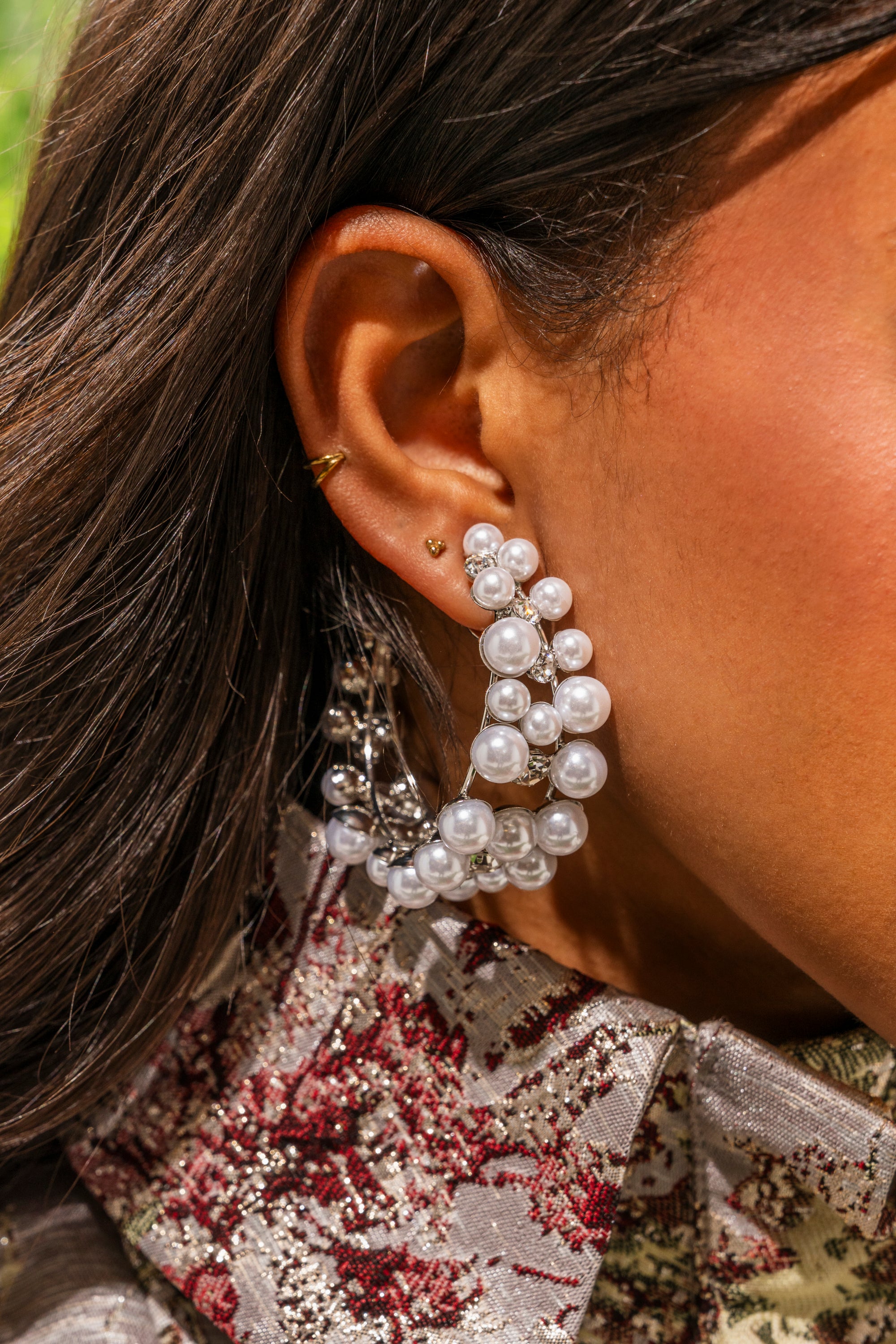 A close-up shows a woman wearing the PEARL PLZ EARRING, a large curved hoop with clustered white pearls and silver beads. She also has two small gold studs, a gold hoop earring, and a floral-patterned outfit. 1