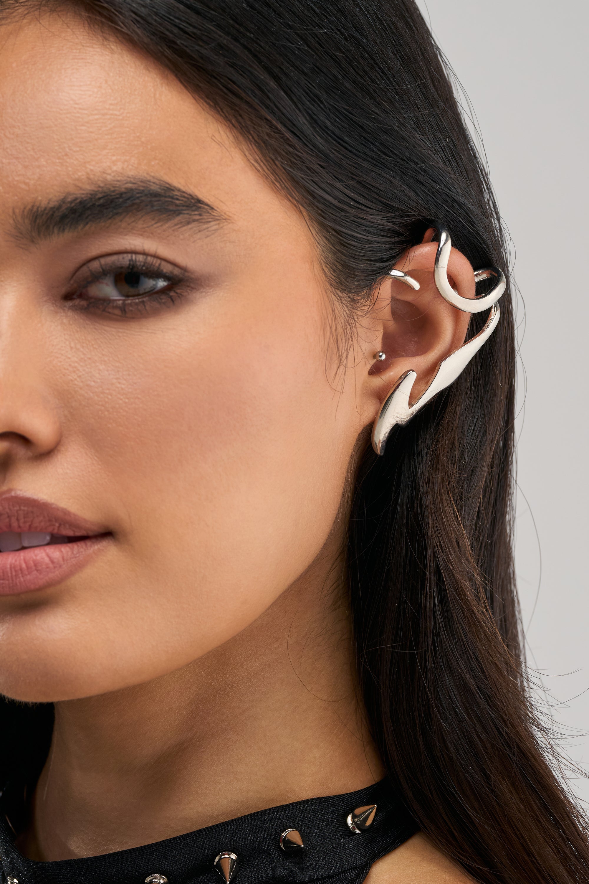 Close-up of a person wearing the E-GIRL EAR CUFF—a large, abstract silver accessory on their right ear. Its sculptural silhouette brings an avant-garde flair to their look. The person has long dark hair, smooth skin, and a neutral facial expression.